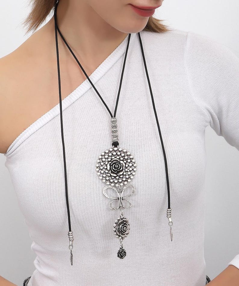May include: A silver necklace with a large, round pendant featuring a floral design. The pendant is attached to a black cord and has a smaller flower charm hanging below it.