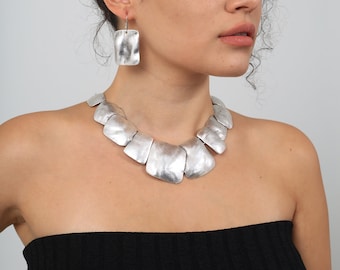 May include: A silver necklace with a geometric design. The necklace is made of multiple connected silver pieces that are slightly curved and have a hammered texture. The necklace is worn by a person with curly hair and a black strapless top.