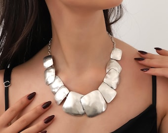 May include: A silver statement necklace with large, irregularly shaped, flat, metal links. The necklace is worn by a person with dark hair and a black top.