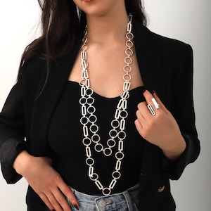 May include: A silver chain necklace with a layered design featuring both square and round links. The necklace is worn by a person with dark hair and a black blazer.