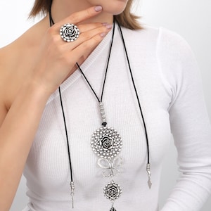 May include: A silver necklace with a large, round pendant featuring a rose design. The pendant is attached to a black cord and has a smaller rose charm hanging below it. The necklace also has a butterfly charm and a small, silver charm.