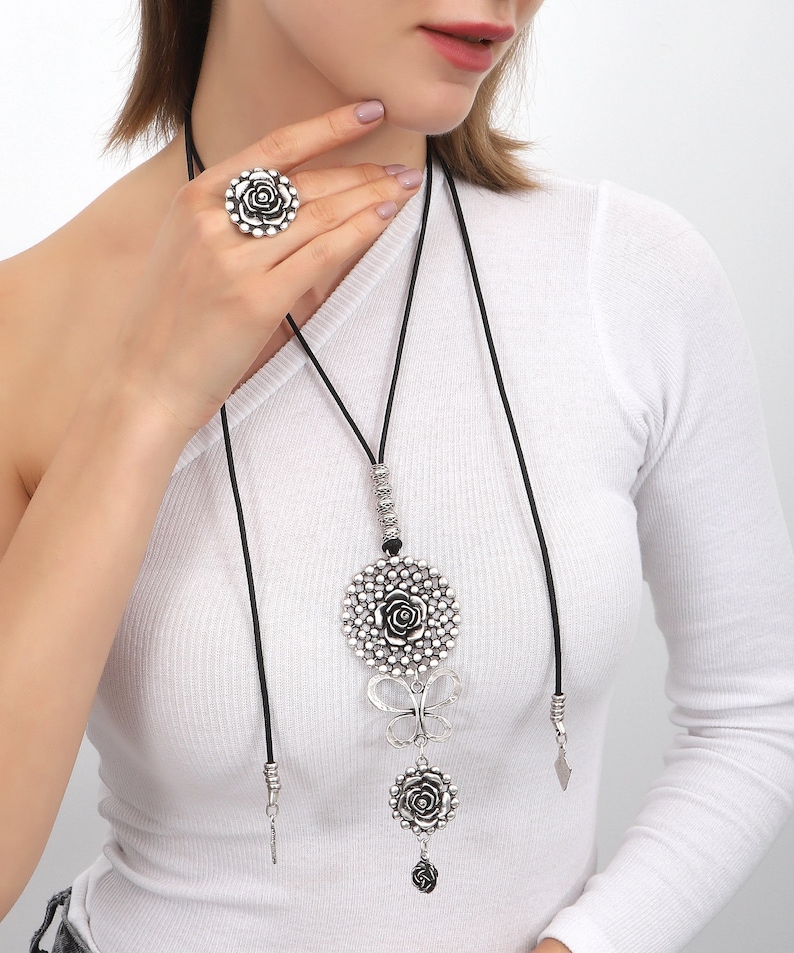 May include: A silver necklace with a large, round pendant featuring a rose design. The pendant is attached to a black cord and has a smaller rose charm hanging below it. The necklace also has a butterfly charm and a small, silver charm.