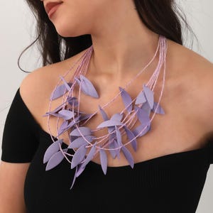 May include: A lavender leaf necklace with multiple strands of pink cord. The leaves are clustered around the neckline, creating a layered effect. The necklace is worn against a black top.