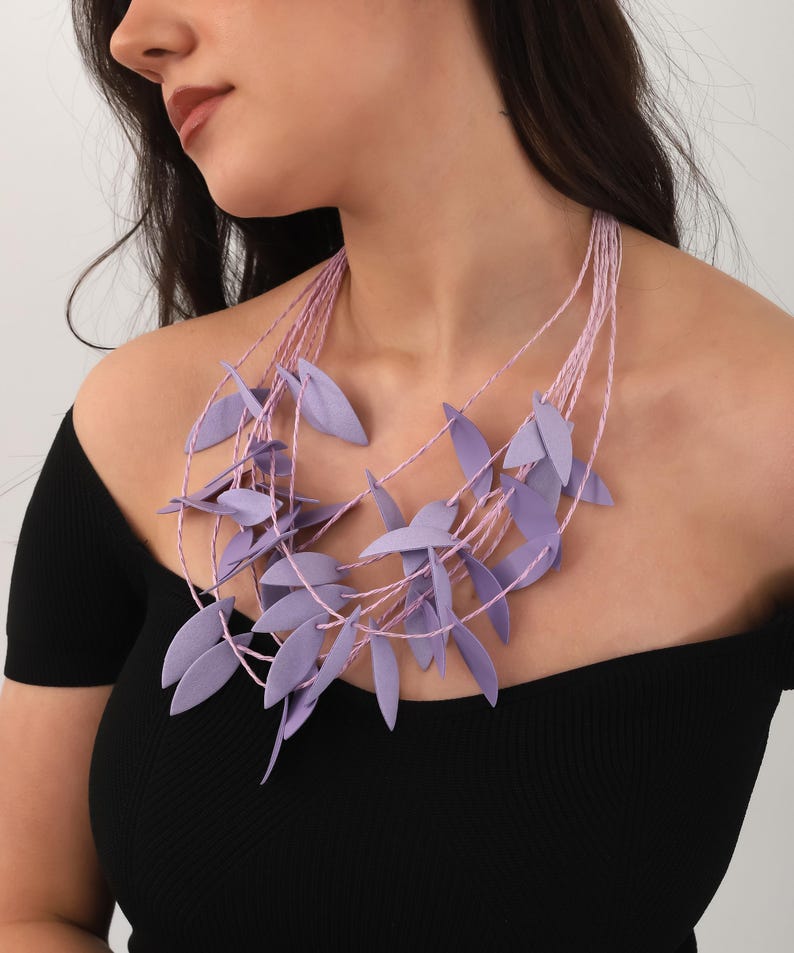 May include: A lavender leaf necklace with multiple strands of pink cord. The leaves are clustered around the neckline, creating a layered effect. The necklace is worn against a black top.