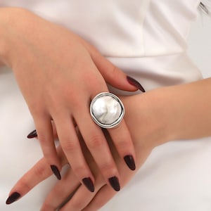May include: A silver ring with a large, round, textured face. The ring is worn on the index finger of a hand with dark red nail polish.
