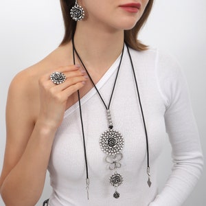 May include: A silver necklace with a large rose pendant and two smaller rose charms. The necklace is on a black cord and has a silver chain with beads. The necklace is worn by a person with a white shirt.