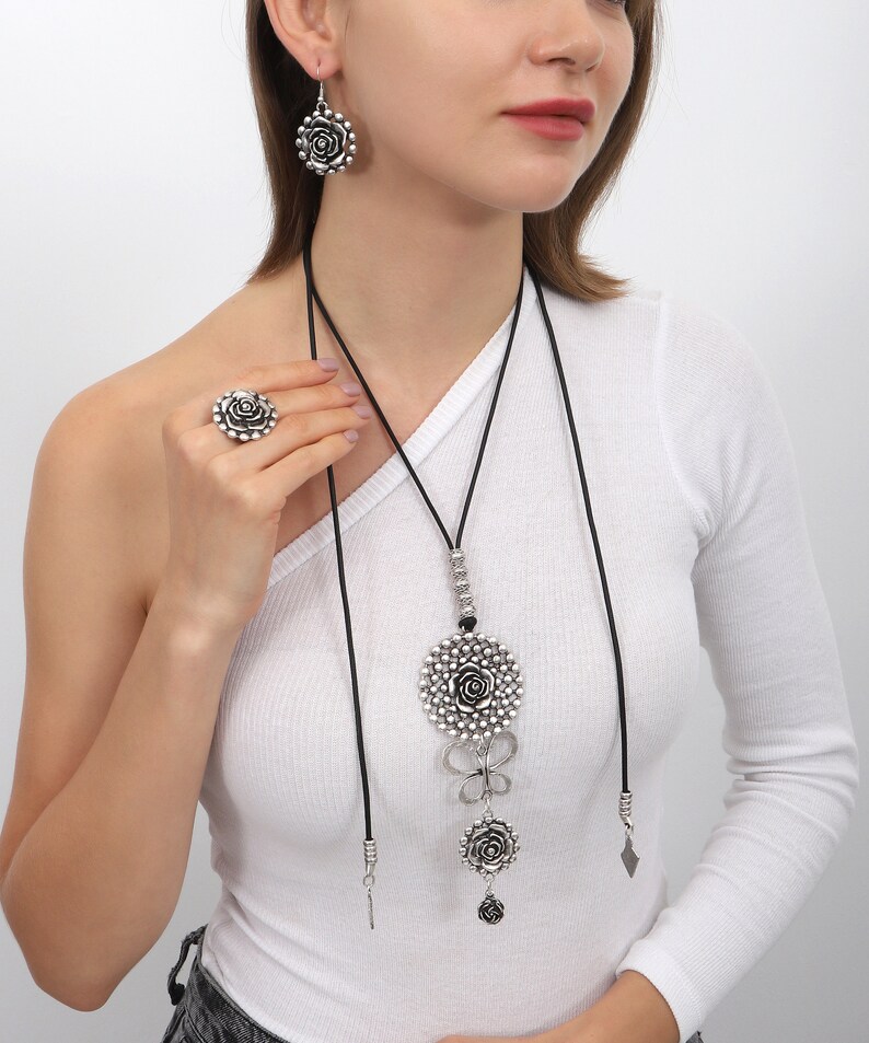 May include: A silver necklace with a large rose pendant and two smaller rose charms. The necklace is on a black cord and has a silver chain with beads. The necklace is worn by a person with a white shirt.
