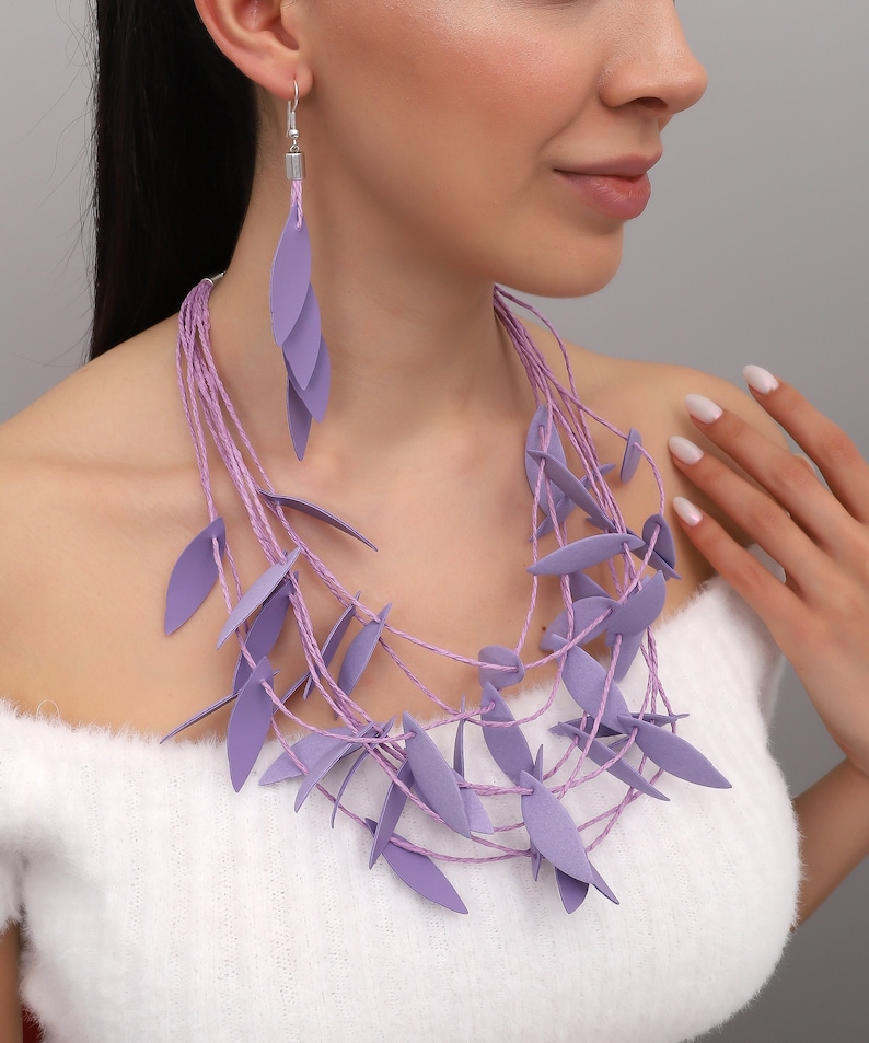 May include: A lavender necklace and matching earrings set. The necklace features multiple strands of lavender-colored cord adorned with leaf-shaped pendants. The earrings have a similar design, with three leaf-shaped pendants hanging from a silver hook.