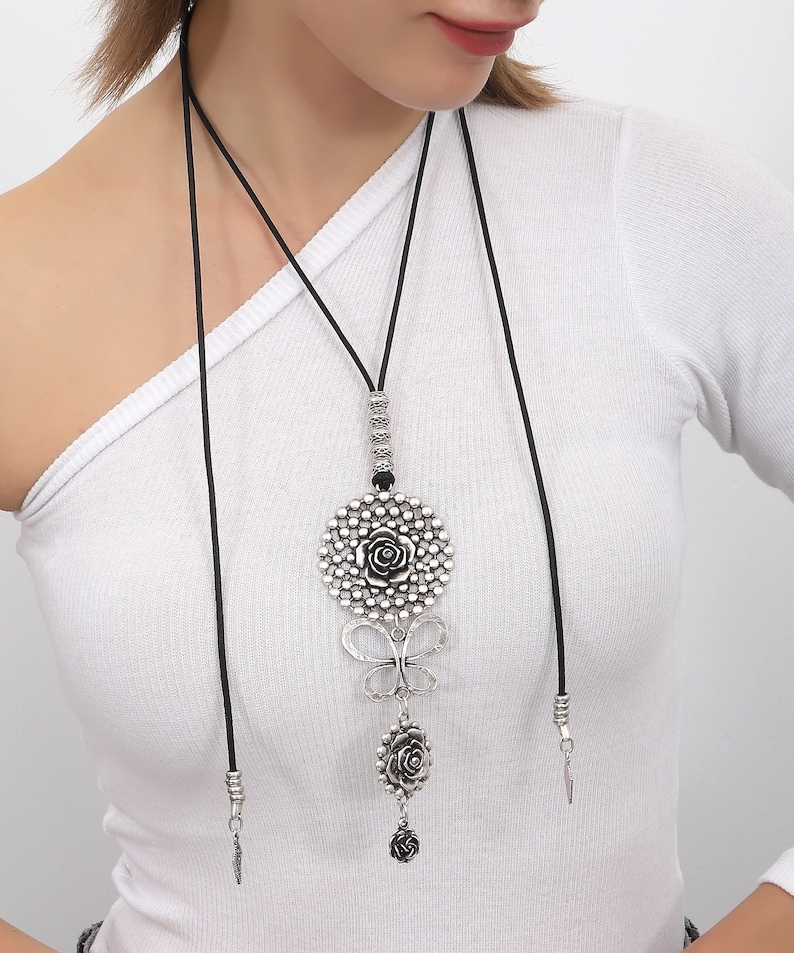 May include: A silver pendant necklace with a large round charm featuring a rose design. The charm is attached to a smaller butterfly charm and a smaller rose charm. The necklace is made of black cord.