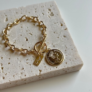 May include: Gold-toned chain bracelet with a toggle clasp. The bracelet features a small rectangular charm and a round coin pendant with a profile image. The bracelet is displayed on a textured, light-colored surface.