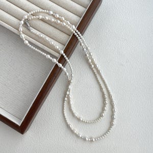 May include: A white pearl necklace with three strands of varying lengths. The necklace is made of small, round pearls and is displayed on a wooden box.