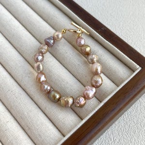May include: A bracelet featuring irregularly shaped, iridescent pearls in shades of pink, gold, and lavender. The pearls are strung together with a gold-colored clasp. The bracelet is displayed on a light beige, textured surface.