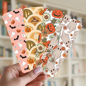 May include: Four rectangular bookmarks featuring Halloween-themed patterns. The first has white ghosts, black bats, and orange pumpkins on a peach background. The second shows pumpkins, candles, and witch hats in scalloped arches. The third displays orange pumpkins and flowers. The fourth features white ghosts and orange flowers.