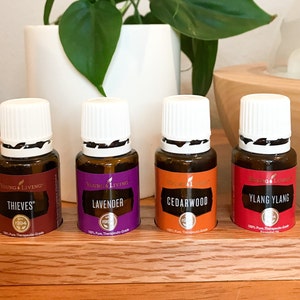 May include: Four small brown glass bottles with white lids and labels. The bottles are labeled "Thieves", "Lavender", "Cedarwood", and "Ylang Ylang". The bottles are arranged in a wooden holder.