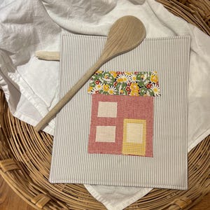 May include: A handmade, striped pot holder with a fabric house applique. The house features a red body, cream-colored windows, and a yellow door. A wooden spoon rests on the pot holder, which is placed on a white cloth.