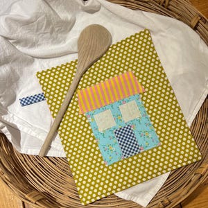 May include: A handmade, square potholder with a house applique, featuring a blue floral print, gingham, and striped roof. The potholder is set on a white cloth with a wooden spoon. The background is a woven basket.