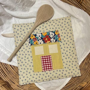 May include: A handmade, square quilted pot holder with a house design. The house features a yellow striped body, a red and white checkered door, and a floral roof. A wooden spoon rests on top of the pot holder, which is set on a white cloth.