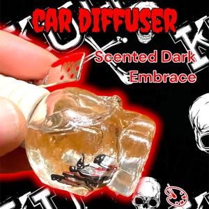 May include: A clear, skull-shaped car air freshener with the text "SKULL HANGING CAR DIFFUSER" in red. The air freshener is filled with a dark liquid and has a small, decorative cleaver. The scent is "Scented Dark Embrace."