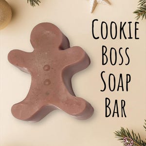 Puede incluir: Una pastilla de jabón marrón con forma de hombre de jengibre. El jabón tiene tres pequeñas hendiduras circulares y el texto "COOKIE BOSS SOAP BAR" en negro. El fondo incluye adornos dorados, una estrella y ramas de hoja perenne.