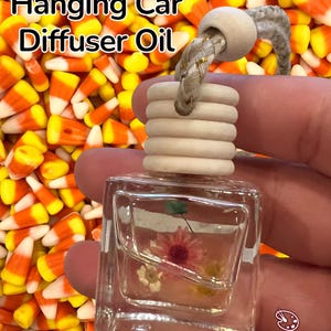 May include: A clear, square-shaped car diffuser oil bottle with a wooden cap and a braided rope. The bottle contains dried flowers and the text "Candy Corn Hanging Car Diffuser Oil". The background is filled with candy corn.
