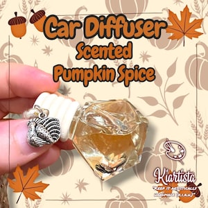 May include: A car air freshener with the text "Car Diffuser Scented Pumpkin Spice." The clear, geometric-shaped bottle contains a light brown liquid. A small silver charm hangs from the white top. The background features fall-themed illustrations.
