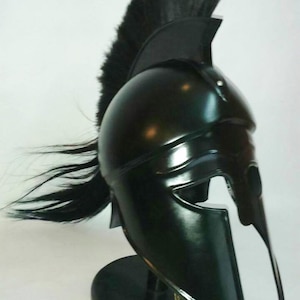 K&ouml;nnte beinhalten: Ein schwarzer spartanischer Helm mit einer schwarzen Rosshaar-Feder auf einem schwarzen St&auml;nder. Der Helm hat einen markanten Wangenschutz und eine erh&ouml;hte Spitze.