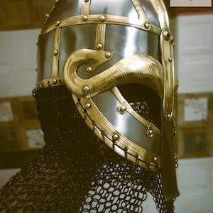 May include: A medieval-style helmet with a polished silver finish and ornate gold-colored accents. The helmet features riveted gold bands, decorative cheek guards, and a chainmail neck guard. The design is reminiscent of historical armor.