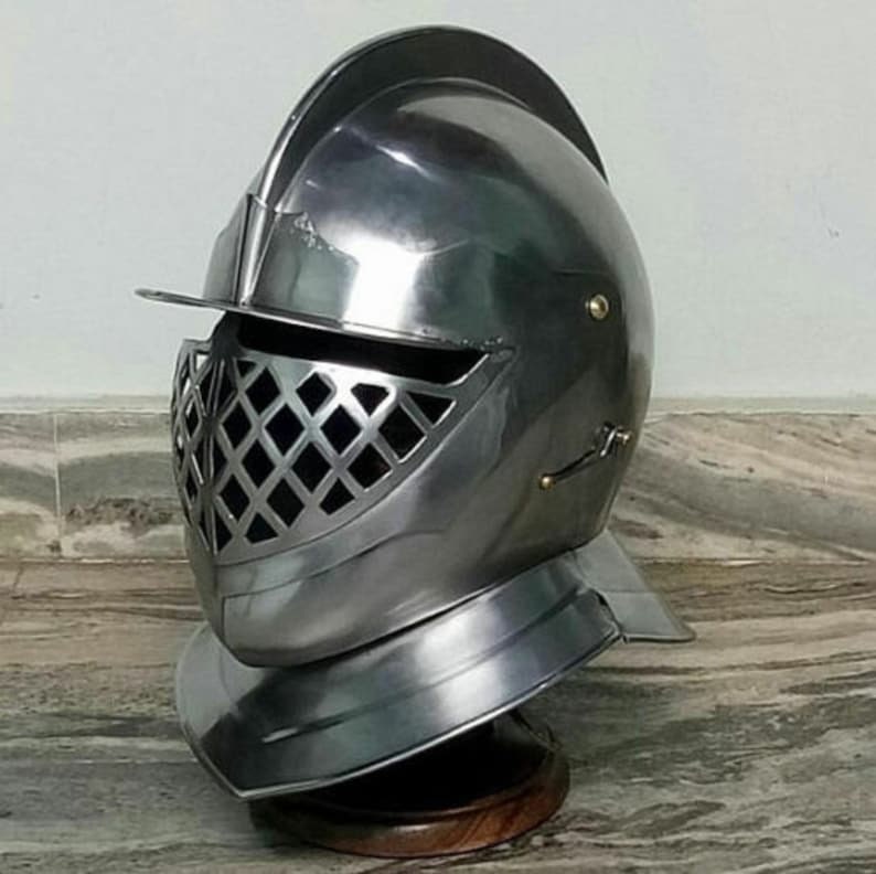 May include: A silver helmet with a visor and a grill-like face mask. The helmet has a rounded top and a pointed chin guard. The helmet is on a wooden stand.