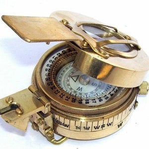May include: A brass compass with a folding lid and a magnifying glass. The compass face has a white background with black markings and a red needle. The compass is marked with numbers from 1 to 360.
