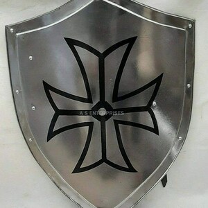 Puede incluir: Un escudo plateado con un dise&ntilde;o de cruz negra. El escudo tiene una parte superior redondeada y una parte inferior puntiaguda. La cruz es un dise&ntilde;o simple con cuatro brazos que tienen la misma longitud. El escudo est&aacute; decorado con peque&ntilde;os remaches de metal alrededor del borde.