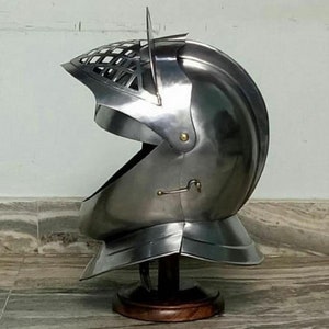May include: A silver helmet with a visor and a decorative grill on the top. The helmet is on a wooden stand.