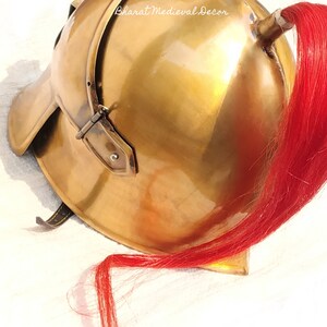 May include: A polished brass helmet with a red plume. The helmet has a rounded top, a curved brim, and a leather strap. The text "Bharat Medieval Decor" is visible on the helmet. This item is likely a historical or costume piece.