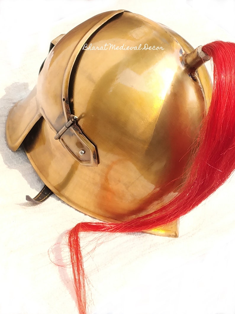May include: A polished brass helmet with a red plume. The helmet has a rounded top, a curved brim, and a leather strap. The text "Bharat Medieval Decor" is visible on the helmet. This item is likely a historical or costume piece.