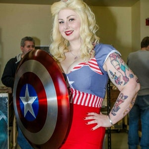 May include: A woman wearing a red, white, and blue costume with a star-shaped brooch on her hair, holding a red, white, and blue Captain America shield. She is smiling and looking at the camera.