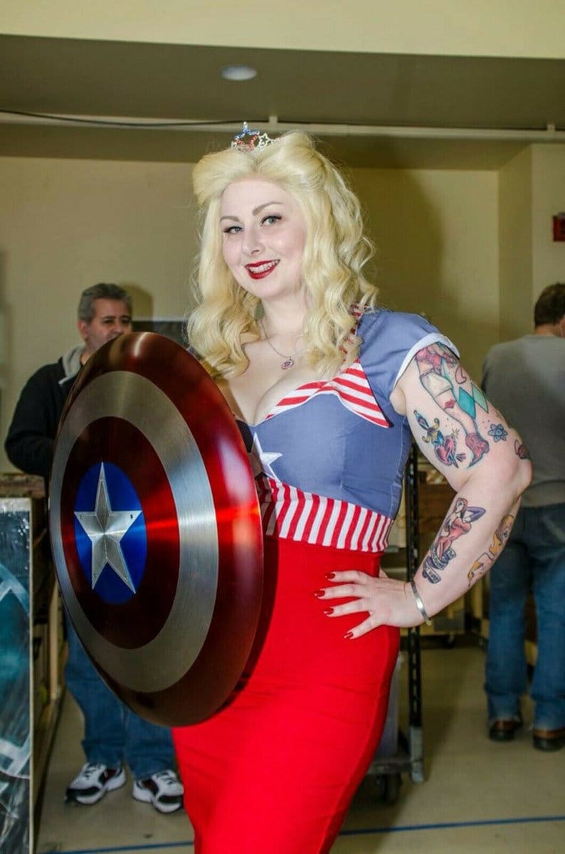 May include: A woman wearing a red, white, and blue costume with a star-shaped brooch on her hair, holding a red, white, and blue Captain America shield. She is smiling and looking at the camera.