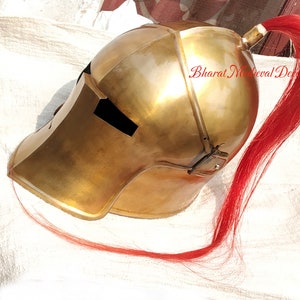 May include: A brass-colored helmet with a red plume. The helmet has a rounded top and a visor with a rectangular opening. The red plume extends from the back of the helmet. The text "Bharat Medieval Decor" is visible.