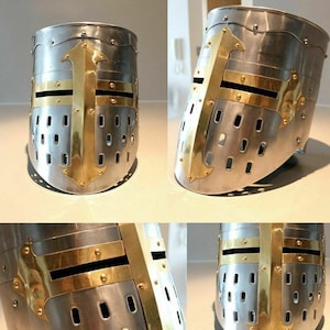 Op de afbeelding: Een middeleeuwse helm met een messing kruis en een vizier. De helm is gemaakt van metaal en heeft een getextureerd oppervlak.