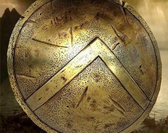May include: A round, gold-colored shield with a raised, triangular design in the center. The shield has a textured surface and appears to be made of metal.