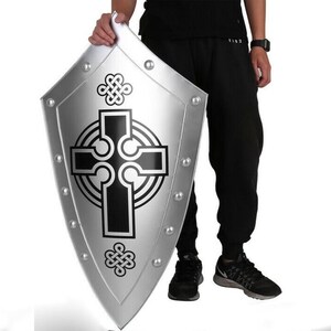 Puede incluir: Un escudo plateado con un dise&ntilde;o de cruz celta negra. El escudo tiene una parte superior redondeada y una parte inferior puntiaguda. Est&aacute; decorado con peque&ntilde;os remaches met&aacute;licos alrededor del borde.