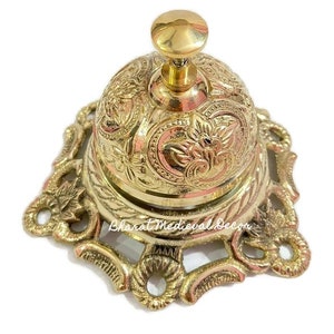 May include: A gold-toned brass bell with an ornate design and a decorative base. The bell has a small knob on top for ringing.