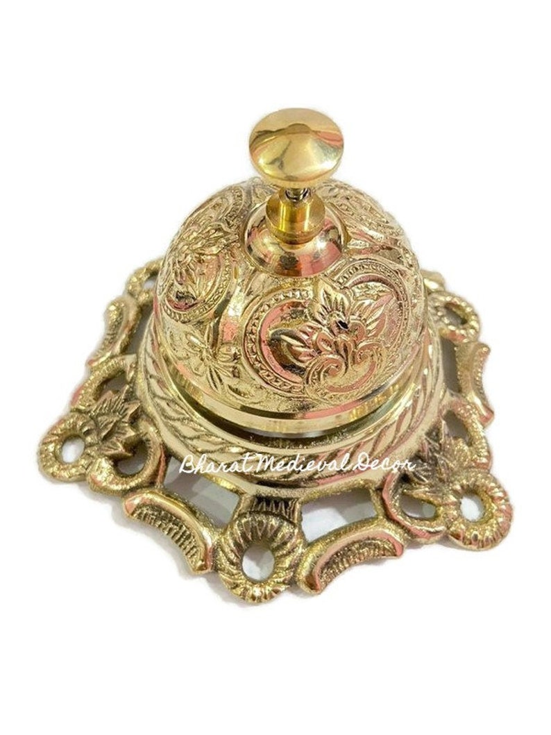 May include: A gold-toned brass bell with an ornate design and a decorative base. The bell has a small knob on top for ringing.