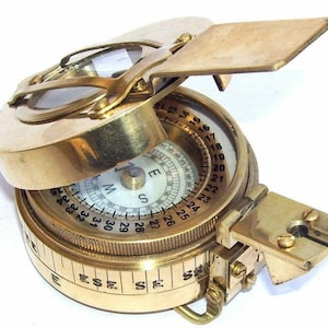 May include: A brass compass with a clear lid and a white face. The compass has a rotating bezel with numbers 1 through 360 and a compass rose with the letters N, E, S, and W. The compass also has a sighting mirror.