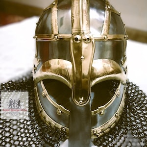 May include: A detailed shot of a Viking helmet, featuring a polished metal finish with brass accents. The helmet has a distinctive nose guard and is surrounded by chainmail. The design is reminiscent of historical armor.