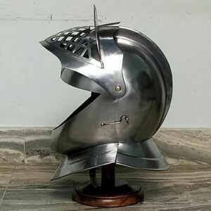 May include: A silver helmet with a visor and a decorative grill on the top. The helmet is on a wooden stand.