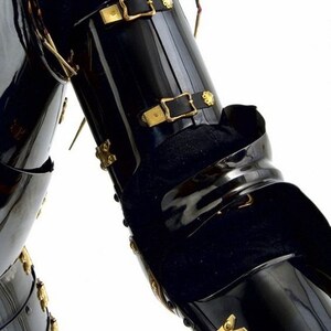 Op de afbeelding: Close-up van een zwart metalen armstuk van een harnas met gouden accenten en leren banden. Het harnas is scharnierend en heeft een decoratief ontwerp.