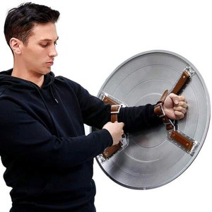 May include: A silver metal shield with brown leather straps. The shield is round and has a raised design in the center. The straps are attached to the shield and can be worn on the arms.