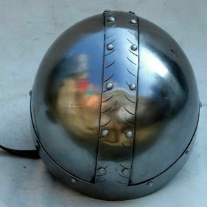 Pu&ograve; includere: Un elmo d'argento con una cupola arrotondata e una fascia decorativa di rivetti metallici. L'elmo ha un cinturino in pelle attaccato al lato.