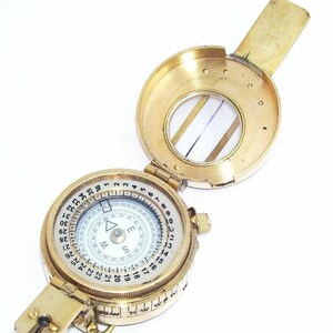 May include: A brass compass with a white face and black markings. The compass has a rotating bezel with numbers 1-360 and a compass rose with the letters N, E, S, and W. The compass is closed and has a small magnifying glass on the top.