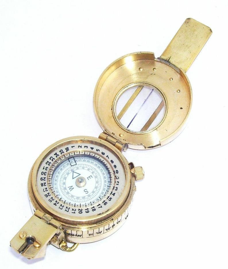 May include: A brass compass with a white face and black markings. The compass has a rotating bezel with numbers 1-360 and a compass rose with the letters N, E, S, and W. The compass is closed and has a small magnifying glass on the top.