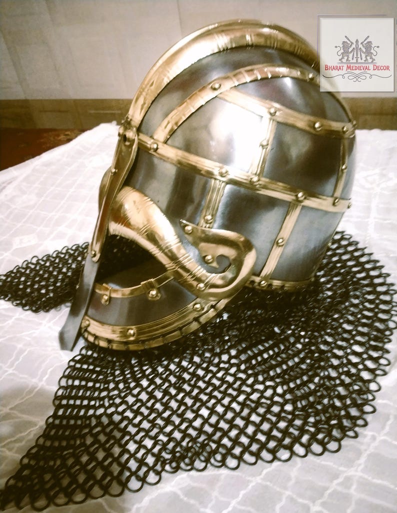 May include: A medieval-style helmet with a polished metal finish and brass accents. The helmet features a curved visor and is adorned with decorative brass strips. A black chainmail coif is beneath the helmet. The logo "BHARAT MEDIEVAL DECOR" is in the upper right corner.